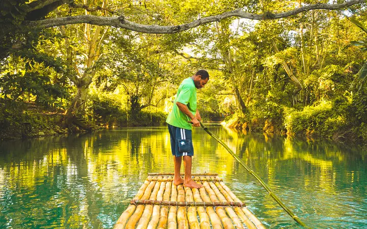 Raft captain poling a bamboo raft on the green Martha Brae River under overhanging trees