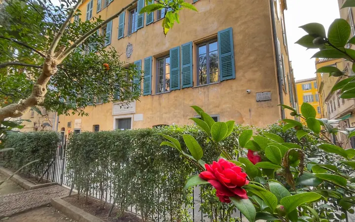 Ochre facade with green shutters of the Maison Bonaparte in Ajaccio, seen across the garden of Place Letizia with red camellias in the foreground