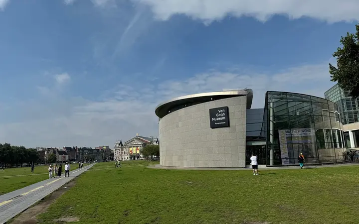 The curved grey exhibition wing and glass entrance hall of the Van Gogh Museum on Museumplein in daylight, with a lawn and footpath in front