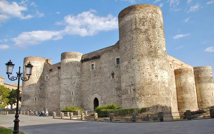The square stone walls and round towers of Castello Ursino in Catania under a blue sky with white clouds, with a street lamp in the foreground