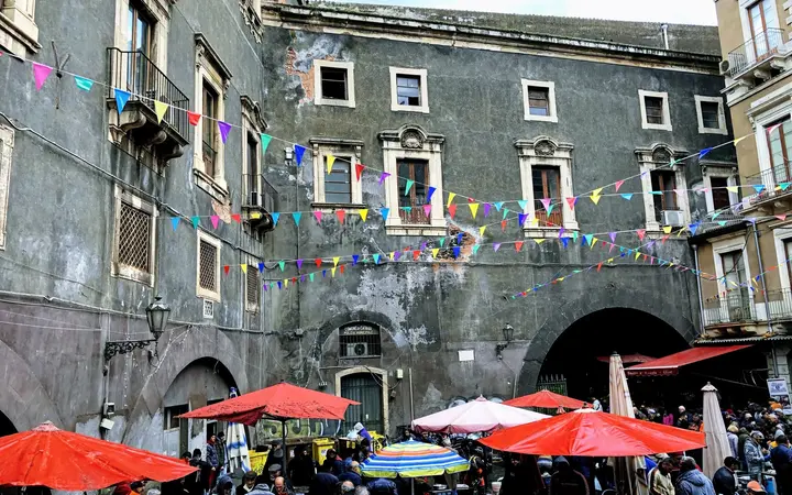 Red and white umbrellas over the stalls of La Pescheria fish market in Piazza Alonzo di Benedetto, with bunting strung across the grey lava-stone buildings