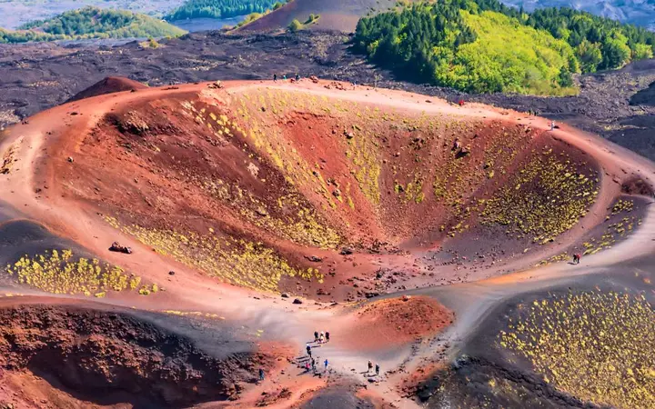 Aerial view of a red and ochre Silvestri crater on Mount Etna, ringed by a footpath with tiny hikers on the rim and pine woods beyond