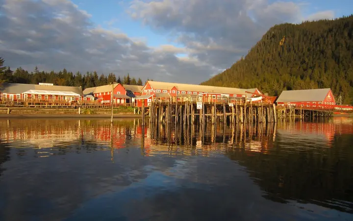 The red historic cannery buildings at Icy Strait Point reflected in calm water, with forested hills behind