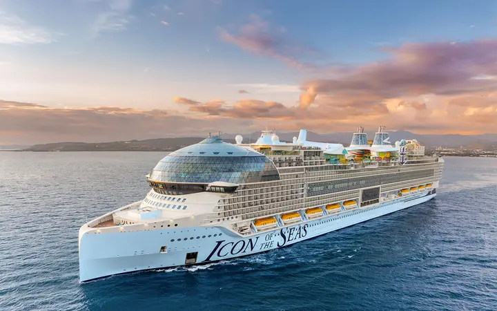Icon of the Seas sailing on open water at sunset, seen from the bow on the port side, with hills on the horizon