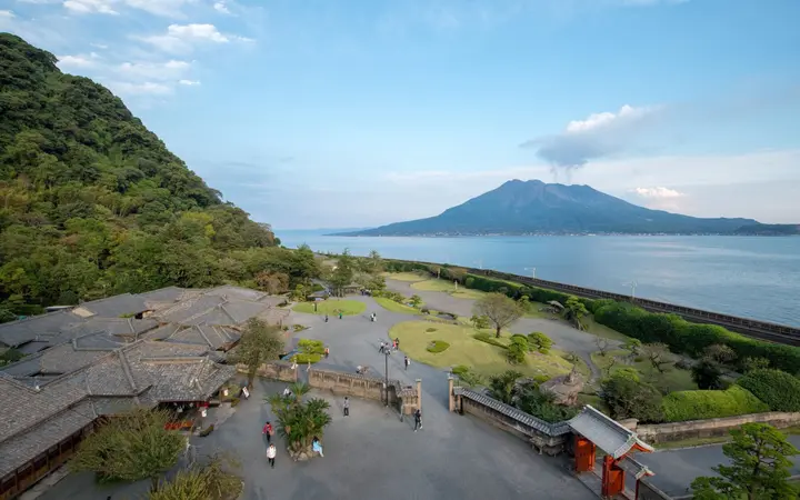 Elevated view of Sengan-en garden and villa roofs with Sakurajima across Kinko Bay under a blue sky.