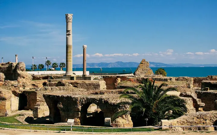 Ruins of the Antonine Baths in Carthage with tall columns, palm trees and the blue Gulf of Tunis behind on a sunny day