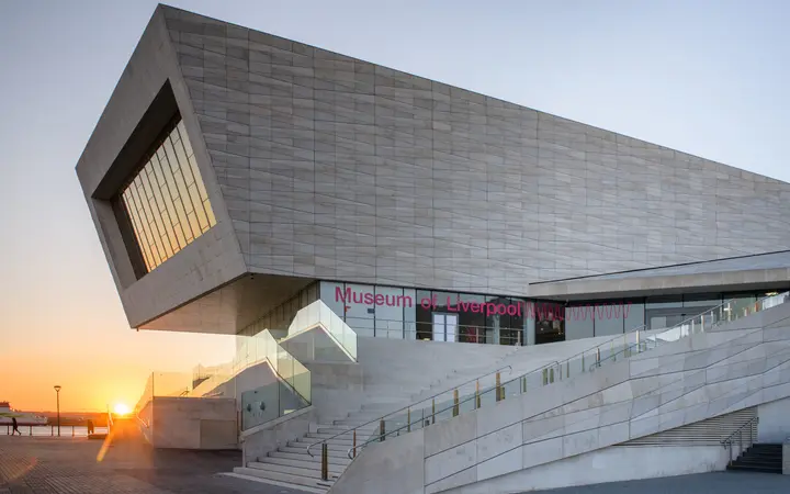 The angular pale-stone Museum of Liverpool building with its large window and outdoor steps on the Pier Head waterfront, sun low on the horizon