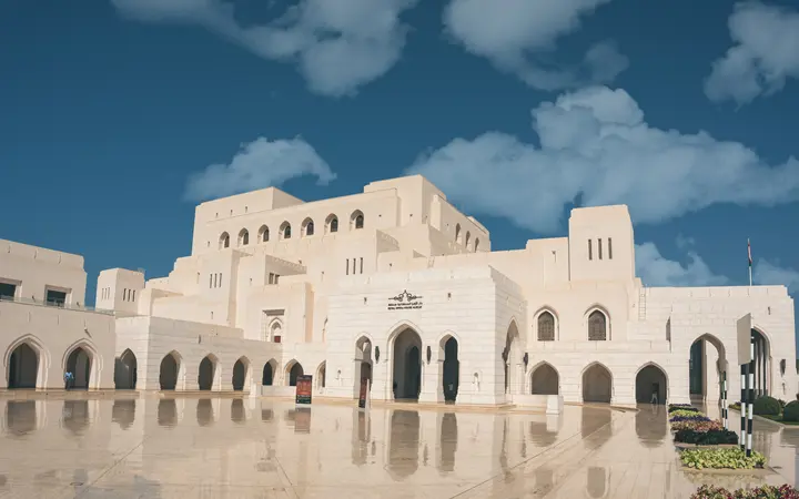 White arched facade of the Royal Opera House Muscat reflected in a forecourt pool.