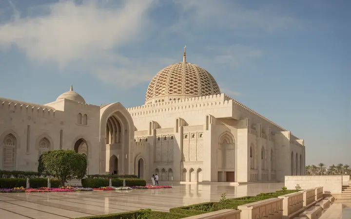 Sultan Qaboos Grand Mosque's sandstone arcades, dome and minaret under a blue sky.