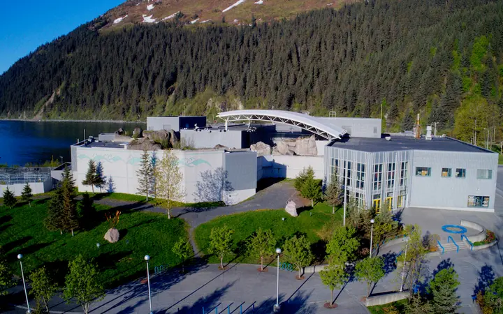 Aerial daylight view of the Alaska SeaLife Center buildings and lawns beside Resurrection Bay, with a forested mountain behind