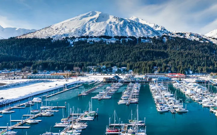 Panorama of the Seward small boat harbor with moored boats below forested, snow-covered mountains under a blue sky