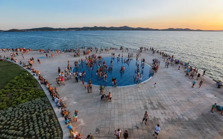 Aerial view of the circular Greeting to the Sun glass installation on the Zadar waterfront at sunset, with visitors and the sea and islands behind